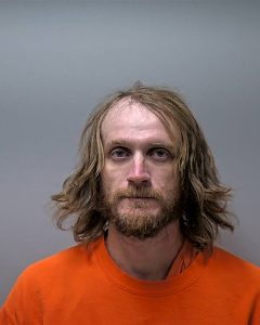 Mugshot of a man with shoulder-length blond hair and beard, wearing an orange prison jumpsuit against a gray background, looking at the camera.