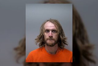 Mugshot of a man with shoulder-length blond hair and beard, wearing an orange prison jumpsuit against a gray background, looking at the camera.