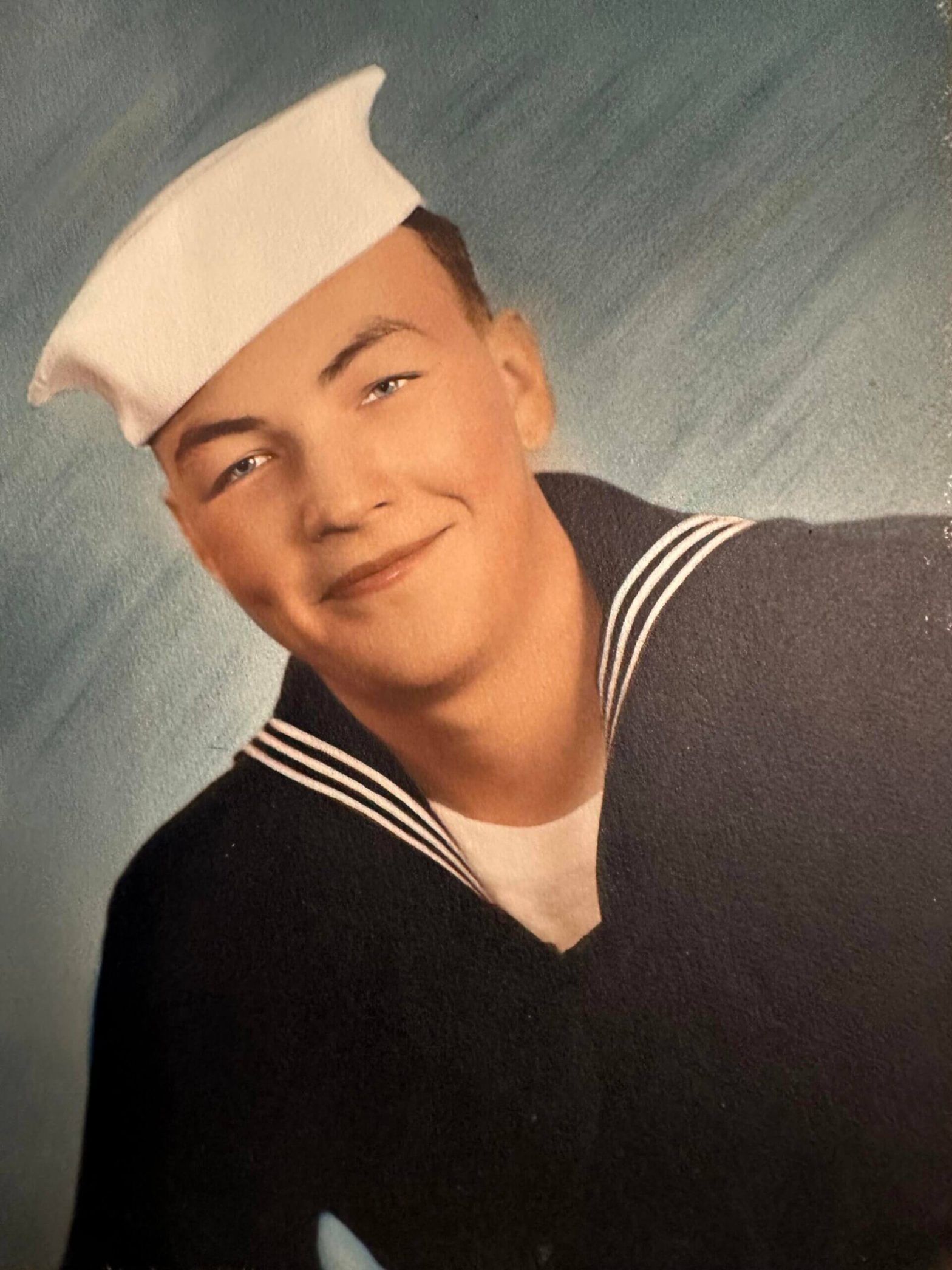 Young man in a navy uniform with a white sailor hat, smiling for a studio portrait.