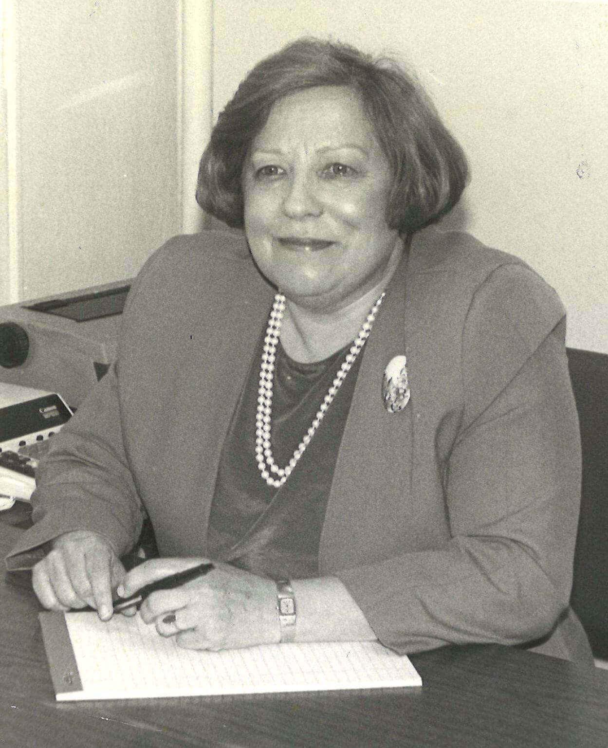 Smiling woman in a blazer with a pearl necklace seated at a desk, signing a document on notepaper.