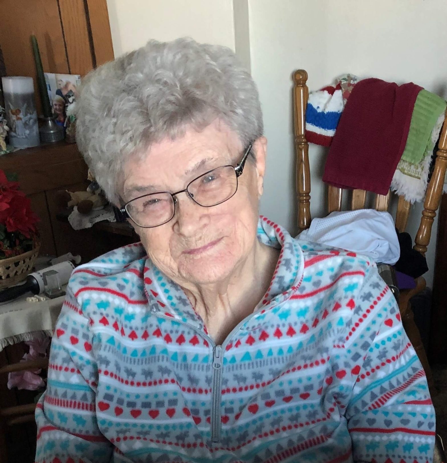 Portrait of an elderly woman with short gray hair and glasses, wearing a pastel patterned zip-up sweater indoors.