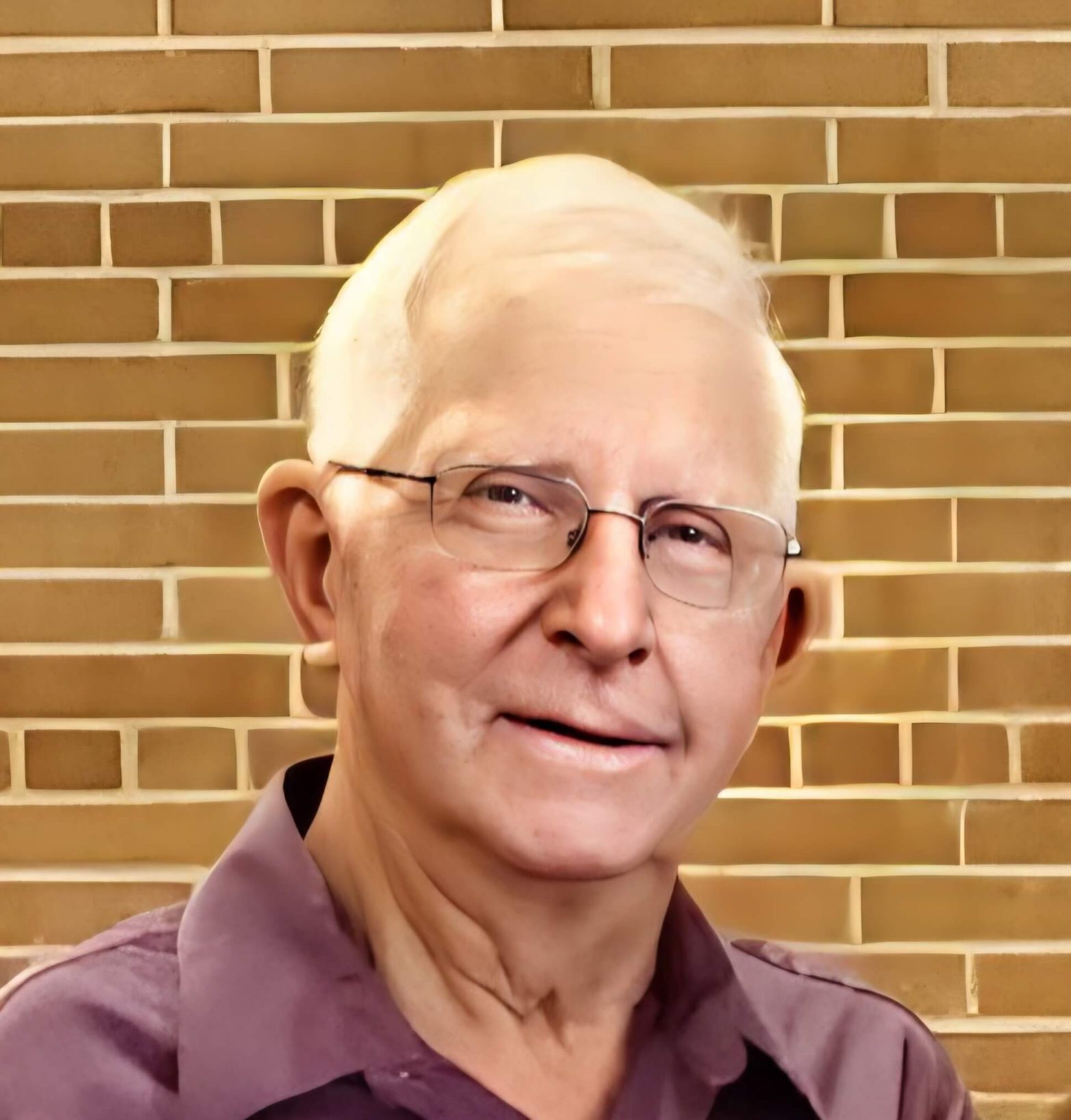 Close-up of a man with glasses, partial face visible, standing in front of a brown brick wall behind him.